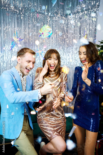 Group of beautiful young people laughing, smiling and holding sparklers over silver shiny background. smartly dressed friends celebrating christmas or new year together, laughing and looking happy