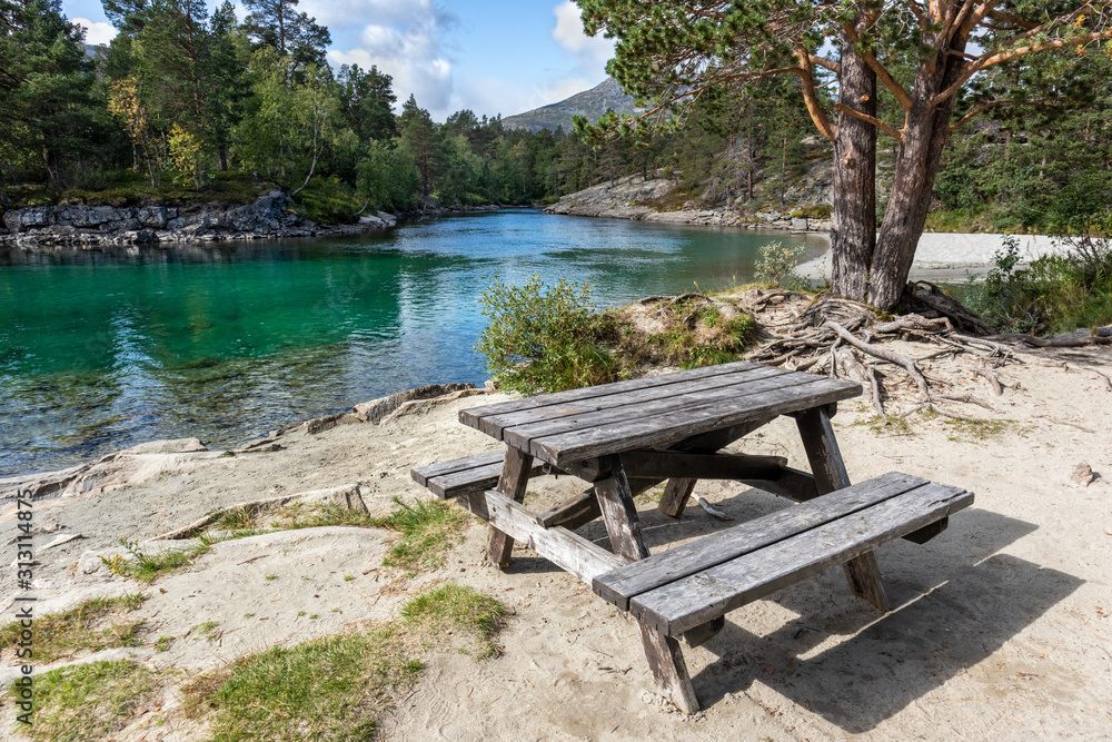 Camping rest area for picnic with wooden table bench on sand by the cold sparkling river in pine forest mountains of Norway
