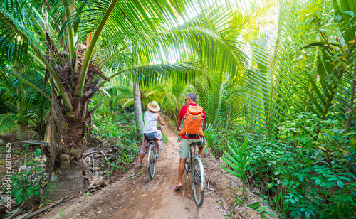 Tourist couple riding bicycle in the Mekong Delta region, Ben Tre, South Vietnam. Woman and man having fun cycling on trail among green tropical woodland and coconut palm trees. Rear view.