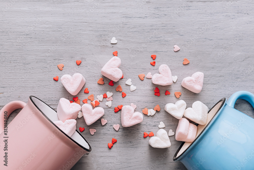 two mugs of blue and pink lie on their side, small candy hearts and marshmallow hearts spilled out