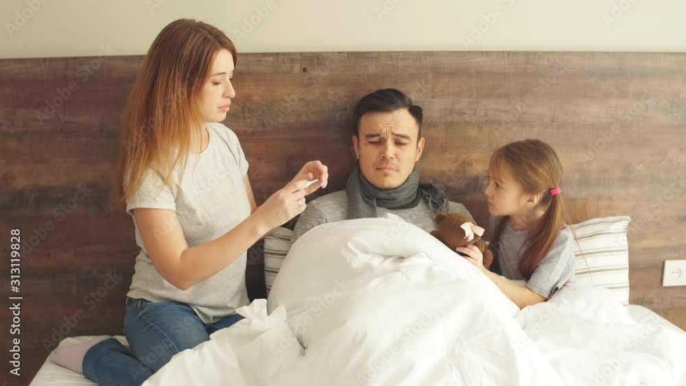 Young caucasian woman measuring temperature of man lying on bed, little child girl sit near him and support Young caucasian woman measuring temperature of man lying on bed, little child girl sit near him and support