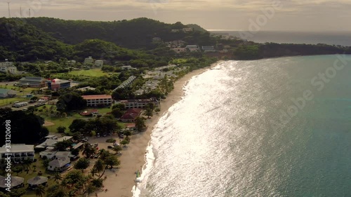 Slow aerial view of the coast of Grenada island, showing residential constructions, calm ocean waves on the sand seashore during dusk, with intense sun reflections on the water