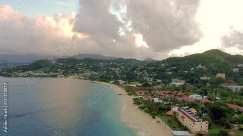 Slow aerial view of the coast of Grenada island, showing residential constructions, calm ocean waves on the sand seashore during dusk, beautiful clouds and lights