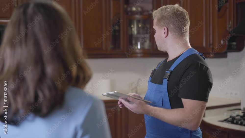Portrait of young handsome Caucasian man in blue robe holding tablet and listening to brunette woman. Unrecognizable lady explaining worker what he has to fix in the house. Service, housekeeping. Portrait of young handsome Caucasian man in blue robe holding tablet and listening to brunette woman. Unrecognizable lady explaining worker what he has to fix in the house. Service, housekeeping.