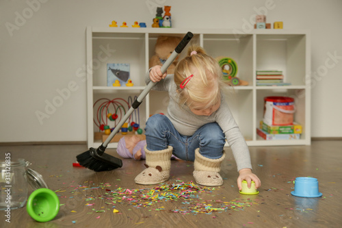 Adorable blonde toddler girl playing with broom, cleaning colorful confetti from the floor photo