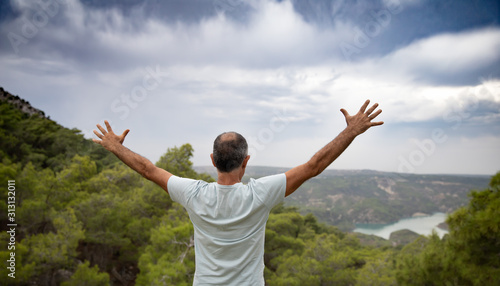 An aged happy man standing on top of a mountain with outstretched arms. Beautiful view of the mountains and sky from the back of man. The concept of freedom and happiness.