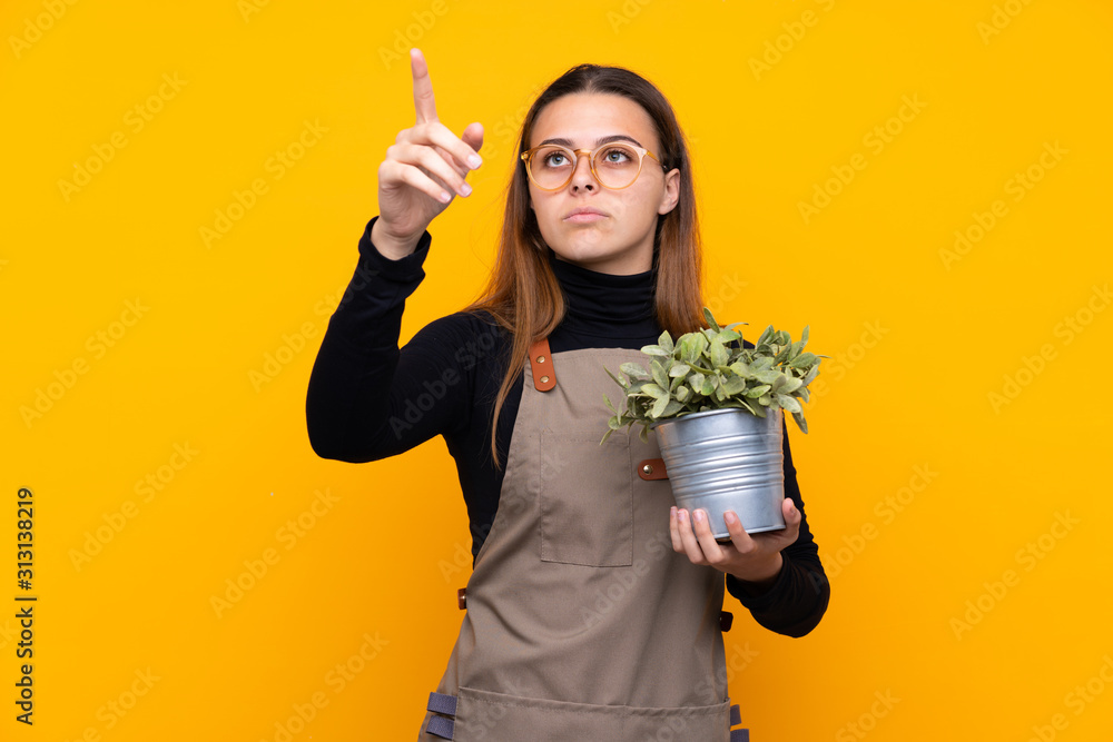 Young gardener girl holding a plant over isolated yellow background touching on transparent screen Young gardener girl holding a plant over isolated yellow background touching on transparent screen