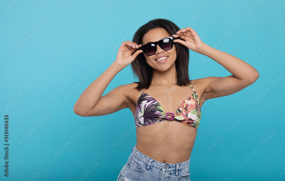 Cheerful african american woman in swimsuit and sunglasses on blue studio background.