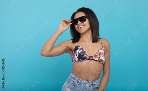 Cheerful african american woman in swimsuit and sunglasses on blue studio background. © lashkhidzetim