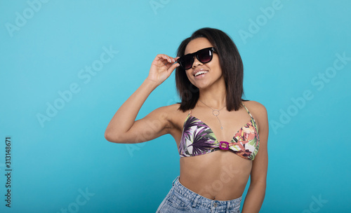 Cheerful african american woman in swimsuit and sunglasses on blue studio background. © lashkhidzetim