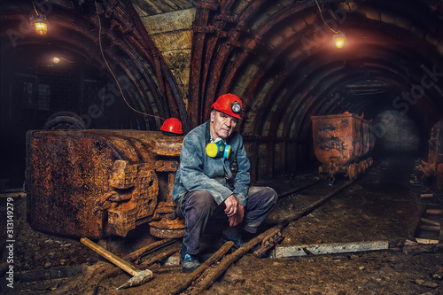 An elderly man dressed in work overalls and a hard hat is standing near the old caravan. Mine worker. A tired miner in a coal mine looks at the light. Work in a coal mine. Portrait of a miner.