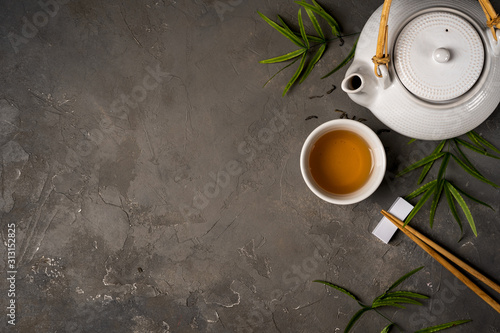 Asian tea concept, cup of tea and teapot surrounded with green tea dry leaves view from above, space for a text on dark stone background