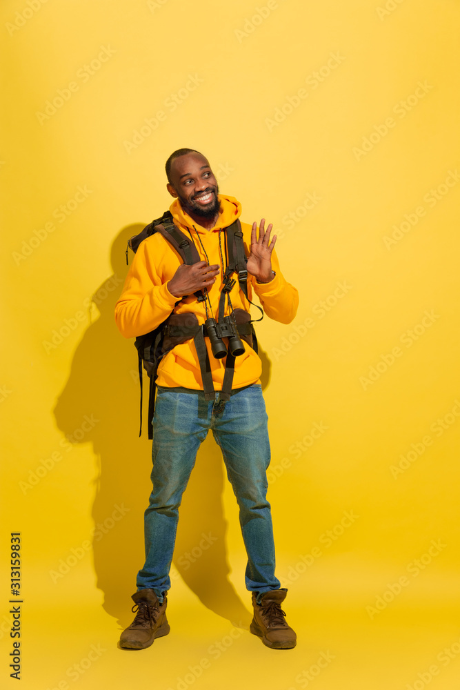 Smiling, looks happy. Portrait of a cheerful young african-american tourist guy with bag and binoculars isolated on yellow studio background. Preparing for traveling. Resort, human emotions, vacation. Smiling, looks happy. Portrait of a cheerful young african-american tourist guy with bag and binoculars isolated on yellow studio background. Preparing for traveling. Resort, human emotions, vacation.