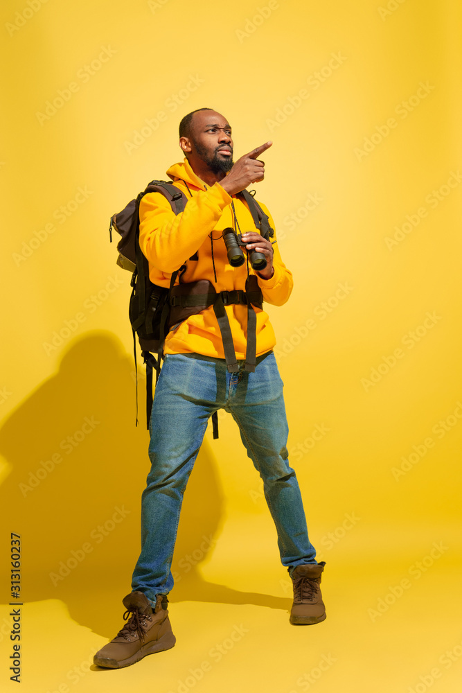 Pointing at side. Portrait of a cheerful young african-american tourist guy with bag and binoculars isolated on yellow studio background. Preparing for traveling. Resort, human emotions, vacation. Pointing at side. Portrait of a cheerful young african-american tourist guy with bag and binoculars isolated on yellow studio background. Preparing for traveling. Resort, human emotions, vacation.