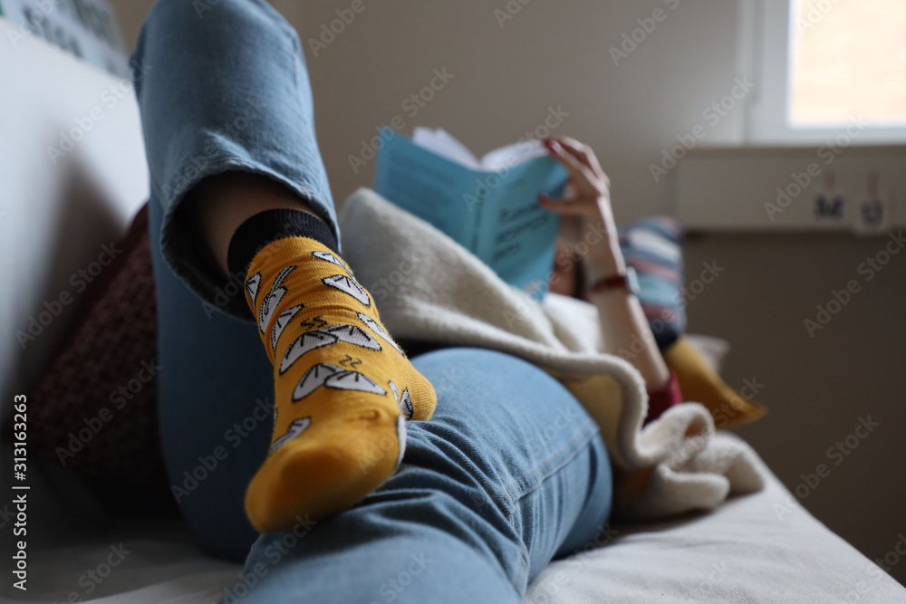 girl in funny socks lies on a sofa reading a book at home, relaxation and comfort - obrazy, fototapety, plakaty 