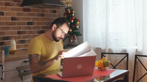 Young caucasian businessman work on laptop at home, hardworking male in casual wear sit in kitchen using modern laptop, with docuents on table photo