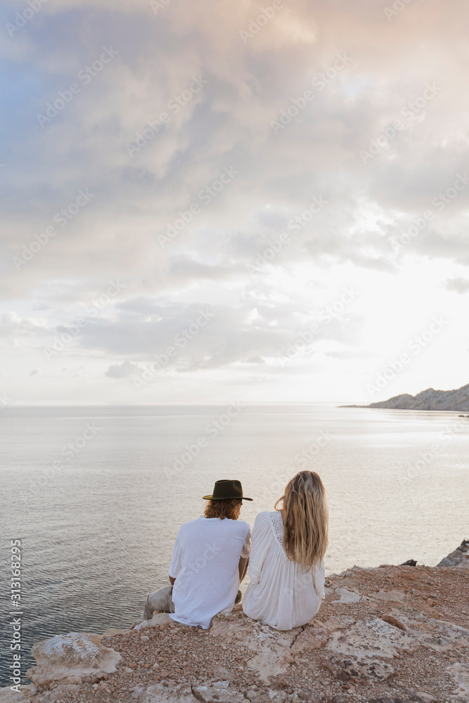 Back view of young couple sitting on rock in front of the sea Back view of young couple sitting on rock in front of the sea