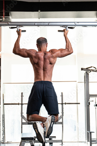 Back view of a strong muscular shirtless mature older bodybuilding athlete with balding gray hair performing a pull-up exercise in a gym