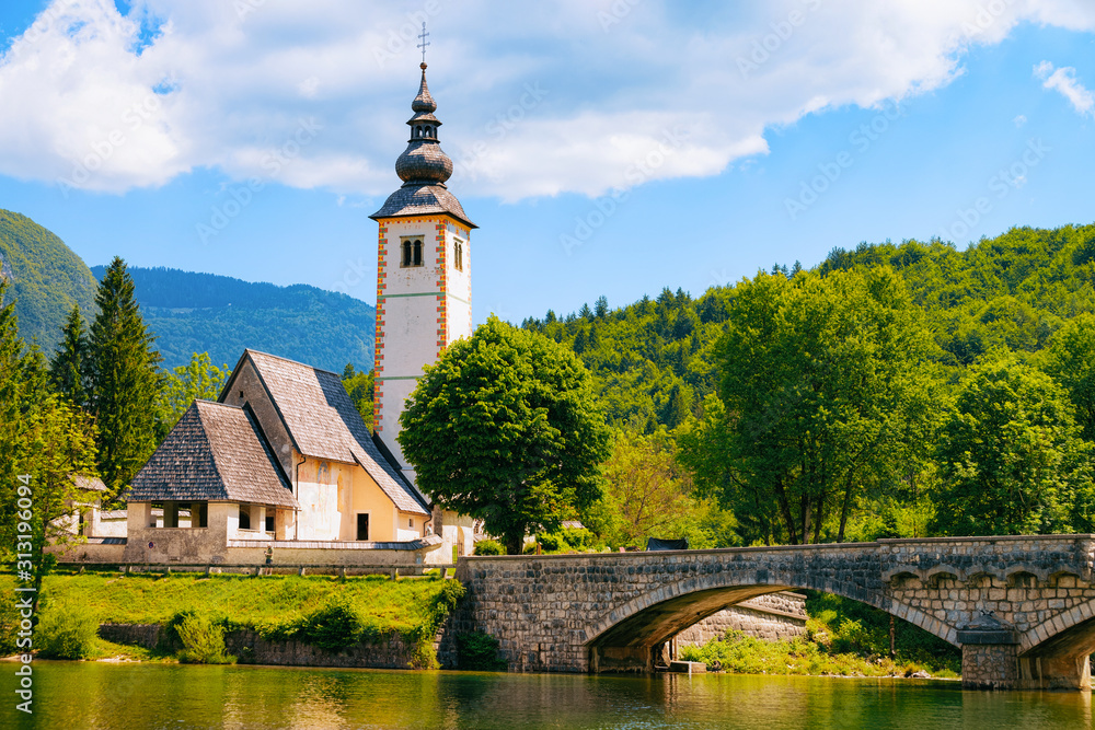 Scenery of Church of St John Baptist on Bohinj Lake at Slovenia. Nature in Slovenija. View of blue sky with clouds. Beautiful landscape in summer. Alpine Travel destination. Julian Alps mountains