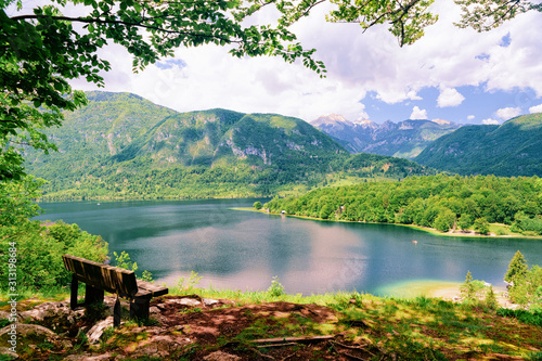 Fototapeta Naklejka Na Ścianę i Meble -  Bench at Bohinj Lake in Slovenia. Nature in Slovenija. Scenery view of green forest and blue water. Beautiful landscape in summer. Alpine Travel destination. Julian Alps mountains on scenic background