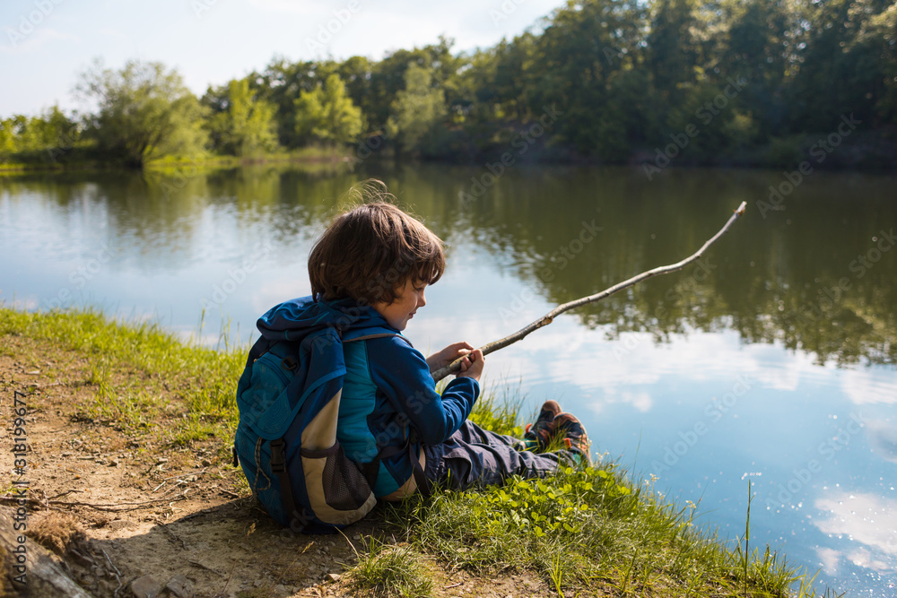 A boy with a backpack sits on the lake with a wooden stick. A boy with a backpack sits on the lake with a wooden stick.