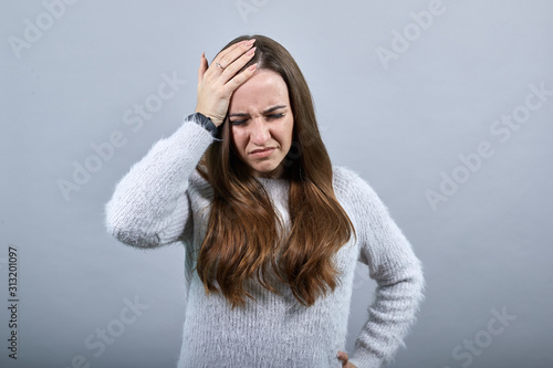 Unhealthy caucasian lady keeping hand on head, having headache, keeping hand on belt wearing blue sweater isolated on gray background in studio. People emotions, lifestyle concept.