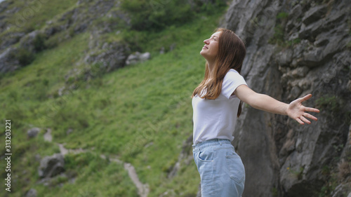 young woman standing on the top of the mountain raises her hands up enjoying the nature.