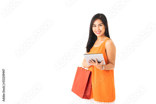 A beautiful Asian young woman wearing orange dress holding shopping bag and a tablet in front of white isolated background.