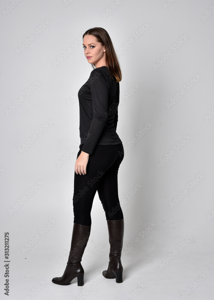 full length portrait of a pretty brunette girl wearing a black shirt leather boots. Standing pose, facing away from the camera, on a grey studio background. full length portrait of a pretty brunette girl wearing a black shirt leather boots. Standing pose, facing away from the camera, on a grey studio background.