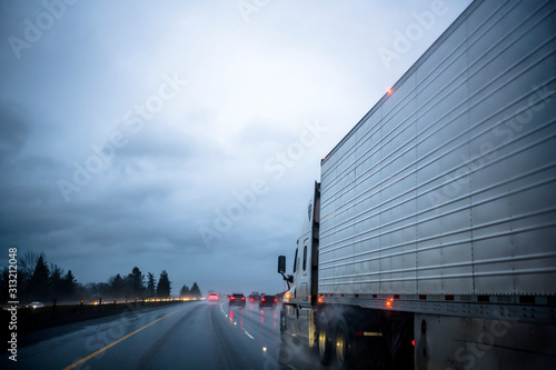 Big rig semi truck transporting frozen cargo in refrigerator semi trailer driving on the raining night highway with reflection light