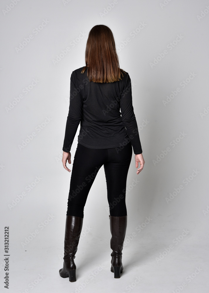 full length portrait of a pretty brunette girl wearing a black shirt leather boots. Standing pose, facing away from the camera, on a grey studio background. full length portrait of a pretty brunette girl wearing a black shirt leather boots. Standing pose, facing away from the camera, on a grey studio background.