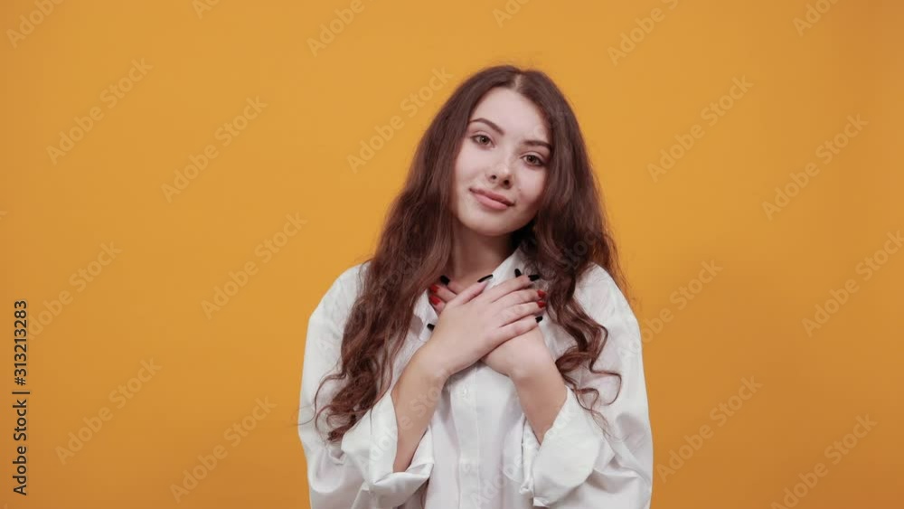 Cheerful caucasian woman in fashion white shirt keeping hands on chest, looking at camera, doing shape of heart isolated on orange background in studio. People sincere emotions, lifestyle concept. Cheerful caucasian woman in fashion white shirt keeping hands on chest, looking at camera, doing shape of heart isolated on orange background in studio. People sincere emotions, lifestyle concept.