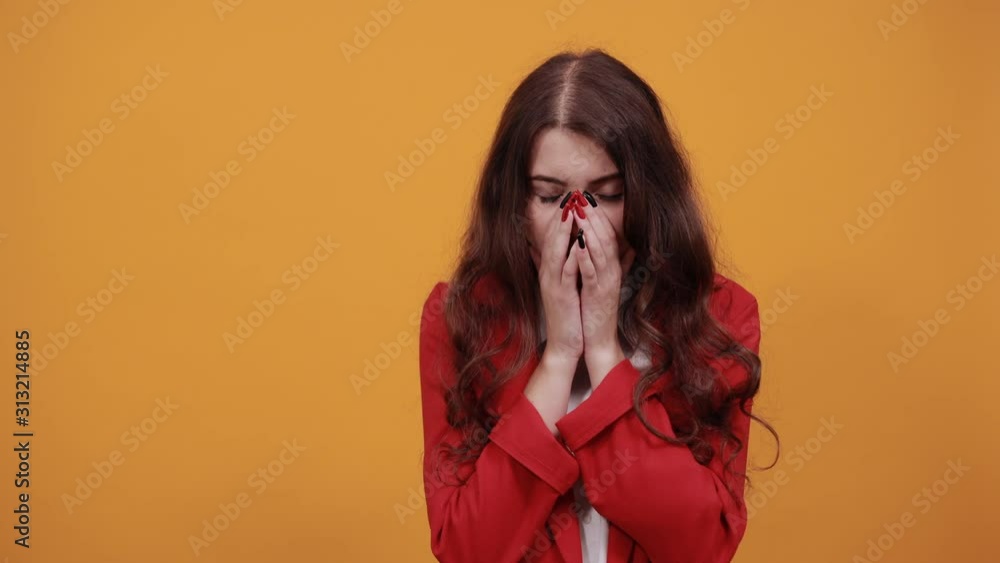 Tired charming brunette young woman in fashion white shirt and red jacket covering nose with hands, crying isolated on orange background in studio. People sincere emotions, lifestyle concept. Tired charming brunette young woman in fashion white shirt and red jacket covering nose with hands, crying isolated on orange background in studio. People sincere emotions, lifestyle concept.