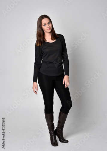 full length portrait of a pretty brunette girl wearing a black shirt leather boots. Standing pose, facing towards the camera, on a grey studio background.