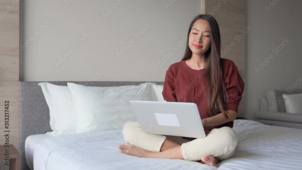 A young Asian woman sits cross-legged on a hotel bed, with a laptop in her lap. A young Asian woman sits cross-legged on a hotel bed, with a laptop in her lap.