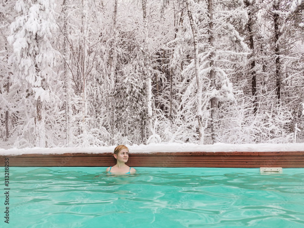 A young fair-haired girl in a blue swimsuit swims in the winter in an open-air pool.  In the background is a snowy forest.  The concept of spa, wellness, Siberia, amazing places for relaxation.