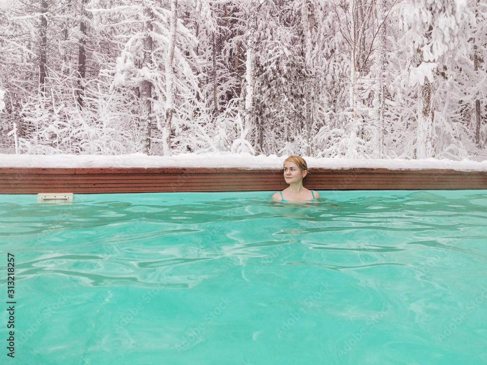 A young fair-haired girl in a blue swimsuit swims in the winter in an open-air pool.  In the background is a snowy forest.  The concept of spa, wellness, Siberia, amazing places for relaxation.