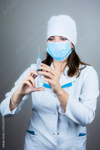 doctor woman in white coat with dark hair in medical blue mask holds syringe in hands on light background photo