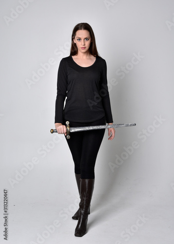 full length portrait of a pretty brunette girl wearing a black shirt and leather boots, holding a sword. Standing pose, holding a sword, on a grey studio background.