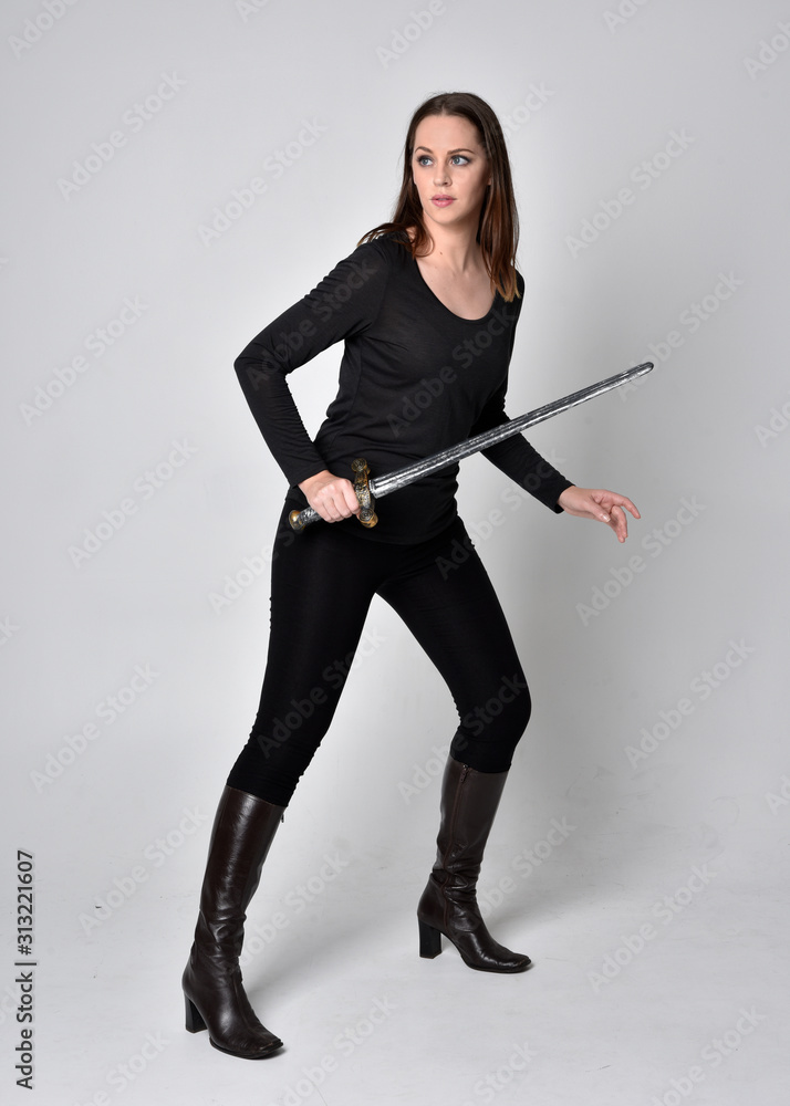 full length portrait of a pretty brunette girl wearing a black shirt and leather boots, holding a sword. Standing pose, holding a sword, on a grey studio background.