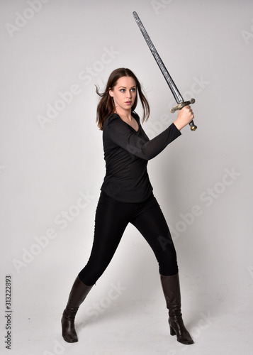 full length portrait of a pretty brunette girl wearing a black shirt and leather boots, holding a sword. Standing pose, holding a sword, on a grey studio background.