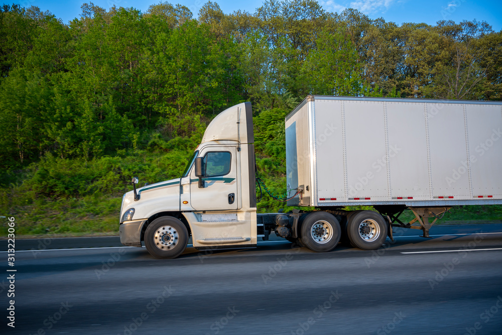 Profile of white big rig day cab semi truck with roof spoiler transporting commercial cargo in dry van semi trailer going on the wide highway with green trees on the roadside