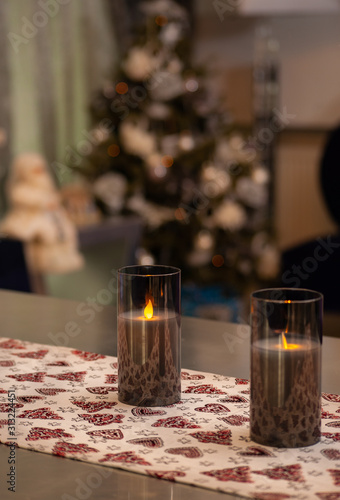 A tablecloth with Christmas pattern on modern designer table with modern candles on it and decorated Christmas tree on the back