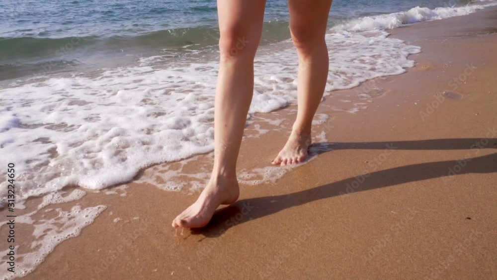 A woman walks along the golden sand of a beach. Female legs walk by the sea. Bare feet of a woman walking along a sandy shore with waves. Summer vacation or vacation. A woman walks along the golden sand of a beach. Female legs walk by the sea. Bare feet of a woman walking along a sandy shore with waves. Summer vacation or vacation.