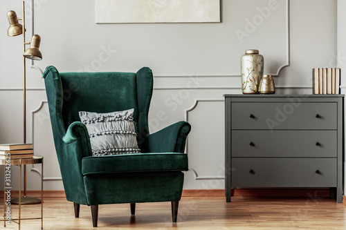 Emerald green wing back chair with pillow in grey living room interior with wooden commode photo