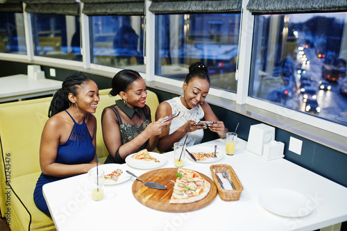 Three african woman in dress posing at restaurant, eating pizza and drink juice, making photos on phone. © AS Photo Family