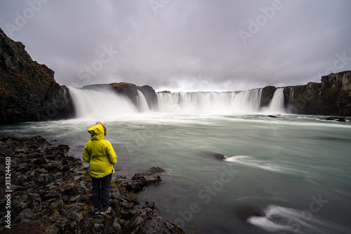 Girl in yellow hiking jacket infront of Godafoss waterfall near Akureyri in the Icelandic highlands. Icelandic and traveling concept.