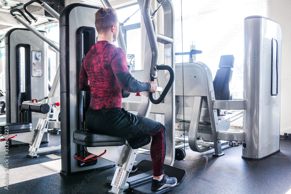 A photo from the back of a sporty muscular man in red and black blouse, engaged in a simulator. A photo from the back of a sporty muscular man in red and black blouse, engaged in a simulator.