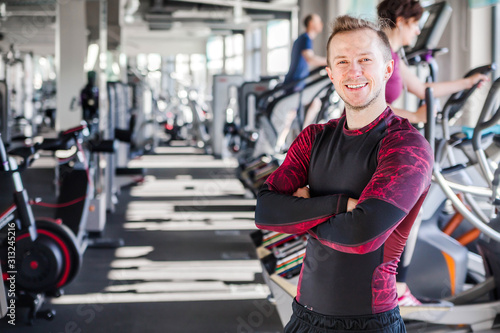 Young man is a fitness instructor, smiling and looking at the camera on the background of the gym. Man crossed his arms