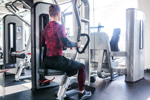 A photo from the back of a sporty muscular man in red and black blouse, engaged in a simulator.